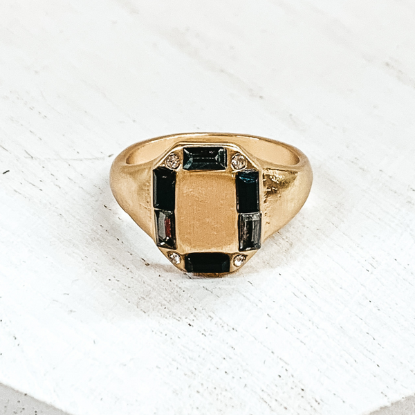 Thick gold band with a rectangle shaped front. The rectangle part includes small, mix of black crystals outlining it. This ring is pictured on a white background.