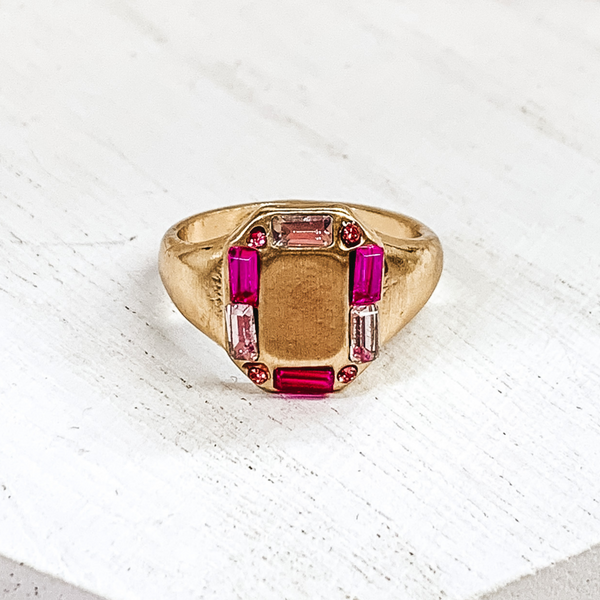 Thick gold band with a rectangle shaped front. The rectangle part includes small, mix of pink crystals outlining it. This ring is pictured on a white background.