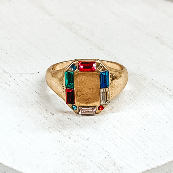 Thick gold band with a rectangle shaped front. The rectangle part includes small, multicolored crystals outlining it. This ring is pictured on a white background. 