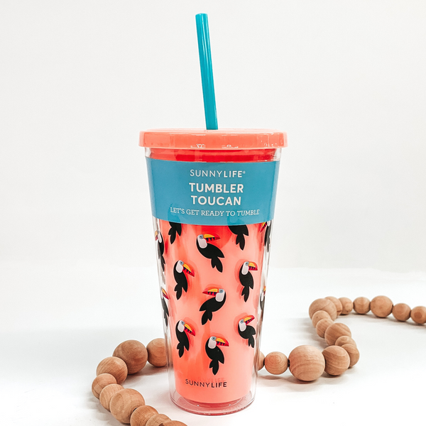 Coral colored double insulated cup. On the outside of the cup there are toucans on the cup. This cup includes a blue straw. This cup is pictured with an blue sticker around the top of the cup with details of the cup. This cup is pictured on a white background with tan beads around it.