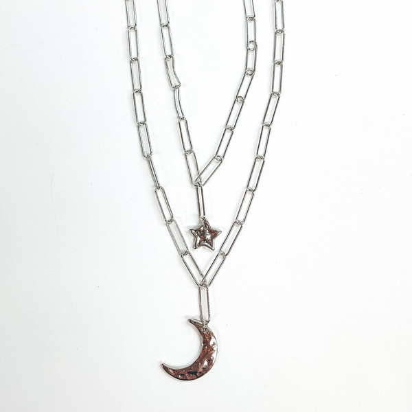 Silver double layered paper clip necklace with two  pendants. The longer layer has a moon pendant and the  shorter layer has a star pendant. This necklace is  taken laying on a white background.