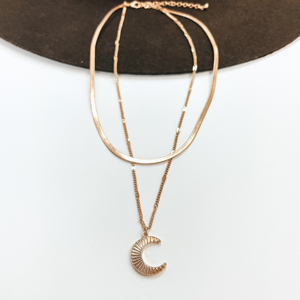 Double layered gold necklace, the smaller necklace  is a snake chain and the bigger one is just a  regular chain. The longer chain has a sunburst  textured moon pendant. Taken on a white background  and laying on a dark brown hat.