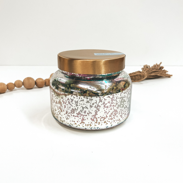 This is a mercury iridescent candle with a gold lid. This candle is taken on a white background with  brown beads in the back as decor.