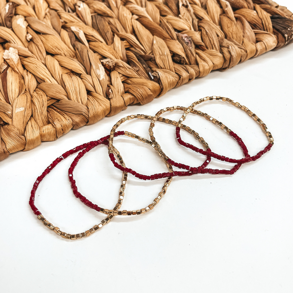 These are five seed beaded bracelets with gold and  burgundy shiny beads. They are taken on a white  background and a brown woven plate in the back as  decor.
