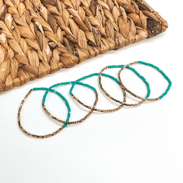 These are five seed beaded bracelets with gold and  turquoise shiny beads. They are taken on a white  background and a brown woven plate in the back as  decor.