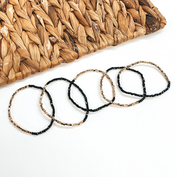 These are five seed beaded bracelets with gold and  black shiny beads. They are taken on a white  background and a brown woven plate in the back as  decor.
