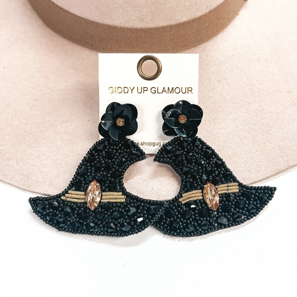 These are witch hat post earrings in black with black crystals and beads, there is gold yarn as a band and with a bronze crystal in the center. The post back is a black flower with a small bronze crystal in the center. These earrings are taken on a beige hat brim and on a white background.