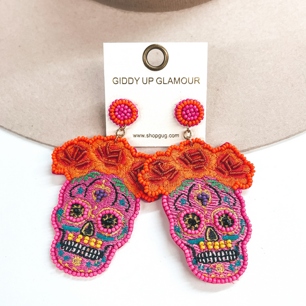These are sugar skull post earrings in fuchsia and other colors. There are orange  flowers in the top with beads and stitching. The skull part has pink beads  around the eyes, yellow beads in the mouth, and black beads in the eyes.  The rest has multicolored stitching all around. These earrings are taken on  a beige hat brim and on a white background.