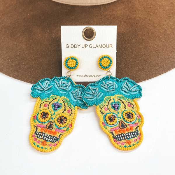 These are sugar skull post earrings in yellow and other colors. There are  turquoise  flowers in the top with beads and stitching. The skull part has yellow beads  around the eyes, orange beads in the mouth, and black beads in the eyes.  The rest has multicolored stitching all around. These earrings are taken on  a dark brown hat brim and on a white background.