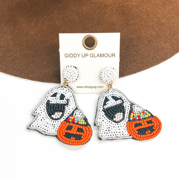 Pictured on a white and brown background are beaded white ghost earrings. These earrings also include a orange pumpkin that has a party hat on. 