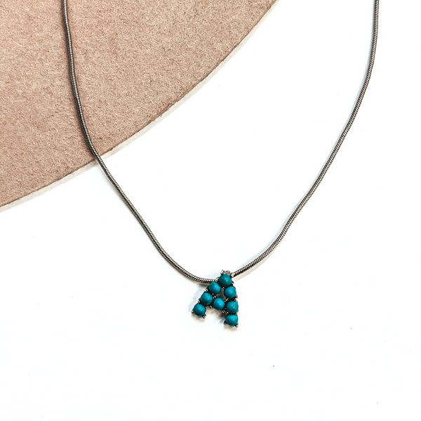 This is a silver, thin, snake chain necklace with the initial A. The  initial is in a silver setting with small turquoise stones. This necklace  is laying on a white background and on a beige felt hat brim.