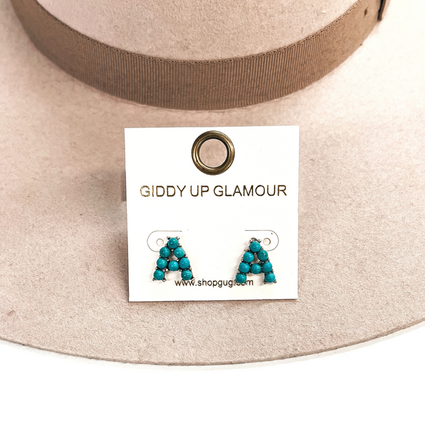 This is a pair of earrings with the initial, A, with small turquoise  stones and in a silver setting. These earrings are placed in a white,  thekitchenapproach card, this is taken on a beige felt hat and on a white  background.