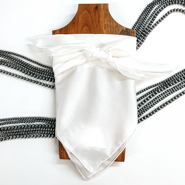 This is a white silk wild rag, this wild rag is placed on a brown necklace board. This wild rag is taken on a white background and with silver Navajo pearls in the back as decor.