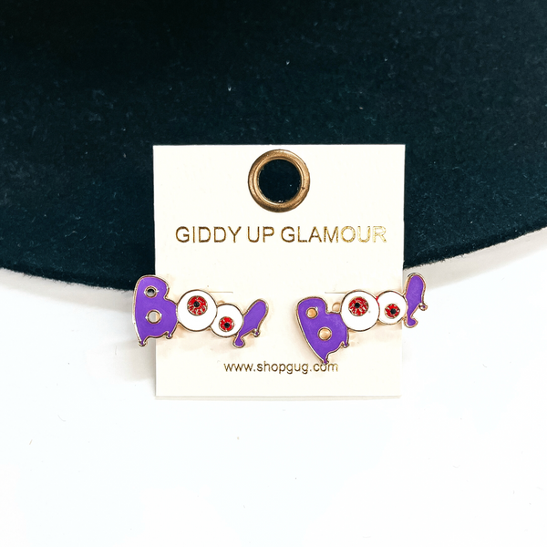 Boo! stud earrings in a purple and white, the B and exclamation point is  purple and the two O's are made to be red eyeballs. These earrings are in  a gold setting. These are taken leaning on a black felt hat brim and on  a white background.