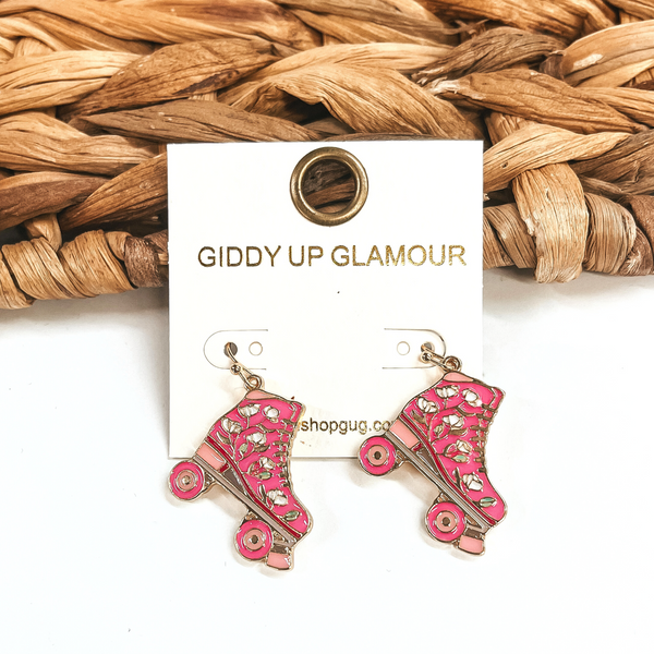 These are pink rollerskate earrings in a gold setting. There is a mix of different  shades of pink; hot pink and light pink. There are white flowers on the rollerskate.  These earrings are taken leaning up against a brown woven slate and on a white  background.