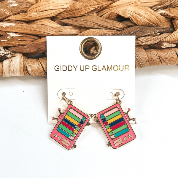These are television shape earrings in pink and multicolor lines on the screen in a  gold setting. There are small hearts in the side of the TV.  These earrings are taken leaning up against a brown woven slate and on a white  background.