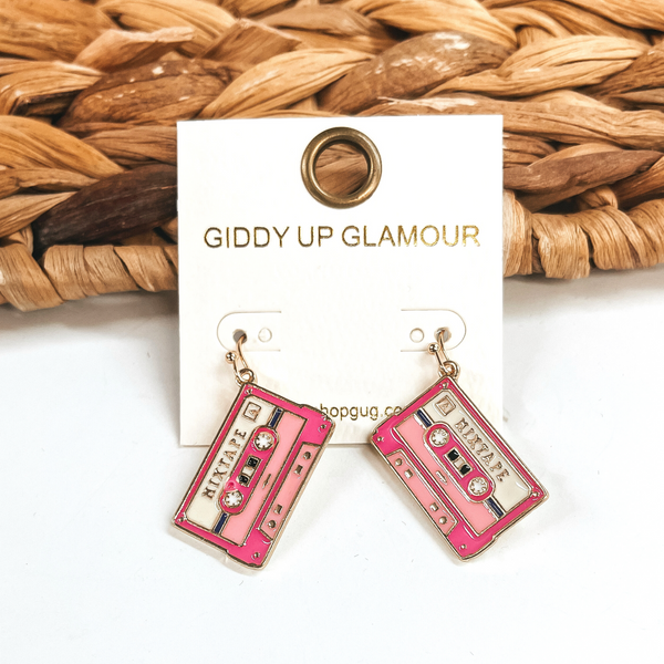 These are pink mixtape shaped earrings in a gold setting with different colors such as  light pink, hot pink, and white. These earrings are taken leaning up against a brown woven slate and on a white  background.