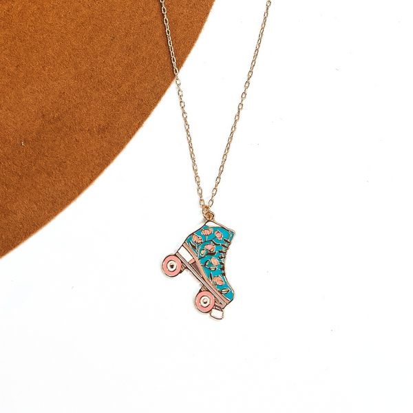 Gold thin chain necklace with a turquoise rollerskate pendant. The turquoise  rollerskate has  light pink floral design in the center. This necklace is taken  laying on a camel-brown, felt hat brim and on a white background.