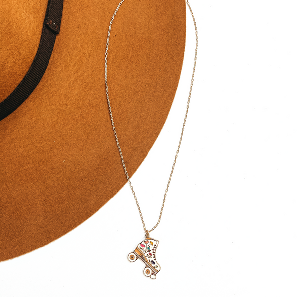 Gold thin chain necklace with a white rollerskate pendant. The white  rollerskate has multicolor floral design in the center such as pink, yellow, blue,  and purple. This necklace is taken  laying on a camel-brown, felt hat brim and on a white background.