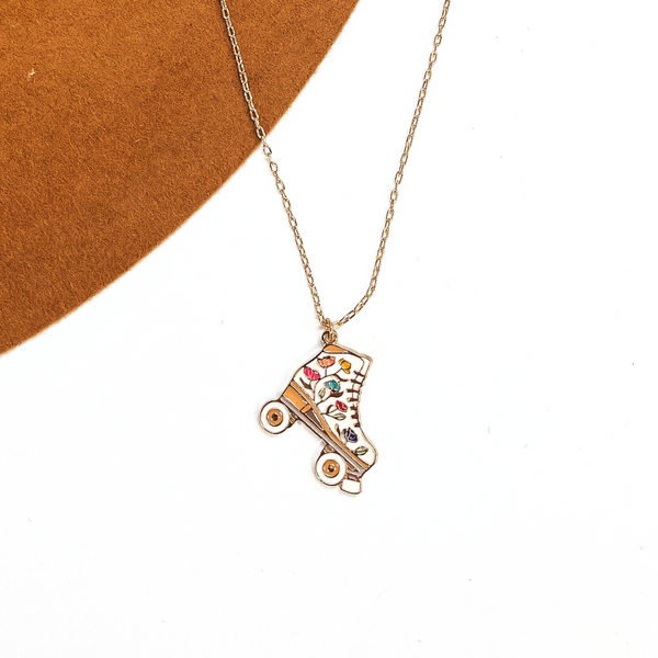 Gold thin chain necklace with a white rollerskate pendant. The white  rollerskate has multicolor floral design in the center such as pink, yellow, blue,  and purple. This necklace is taken  laying on a camel-brown, felt hat brim and on a white background.