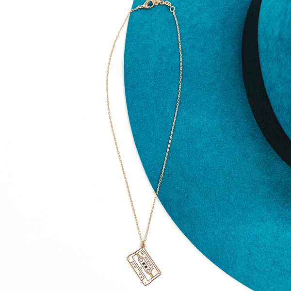 Gold thin chain necklace with an ivory cassette tape pendant. The cassette pendant  has a bit of glitter in the center and other tones of white and tan. This necklace  is laying on a white background and a teal felt hat brim.