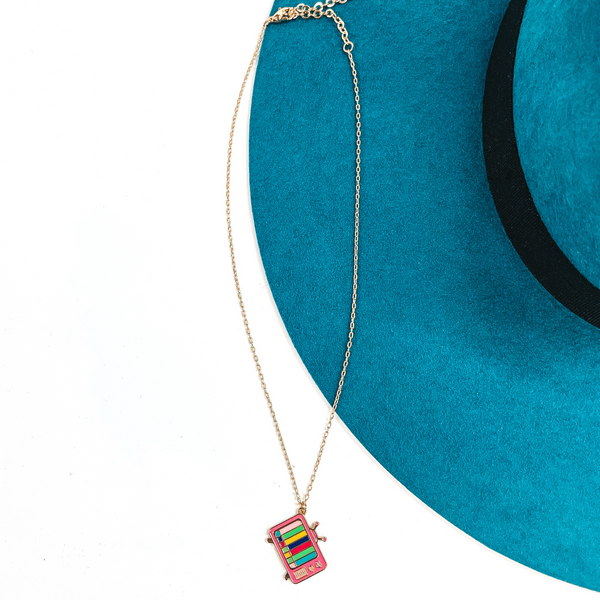 Gold thin chain necklace with an old television set pendant in pink. The tv is on a gold setting has multicolor lines on the screen. This necklace  is laying on a white background and a teal felt hat brim.