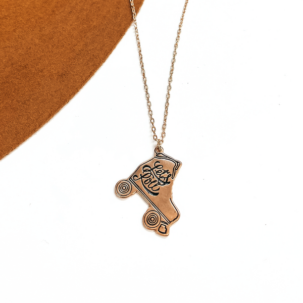 Gold tone thin chain necklace with a gold tone rollerskate pendant that says, 'Let's Roll'. This necklace is taken on a white background and on a camel-brown felt hat brim.